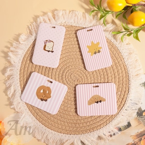 May include: Four rectangular plastic ID badge holders with rounded corners. Each features a light pink background with white patterns and a cartoon food character: a bread slice, a star, a pretzel, and a croissant, all with faces.