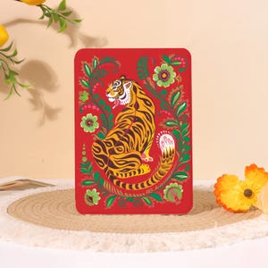 Peut inclure: Un objet décoratif rectangulaire rouge avec une illustration détaillée d'un tigre. Le tigre a des rayures jaunes et noires et est entouré de motifs floraux verts avec des fleurs roses et vertes. L'objet a des coins arrondis.