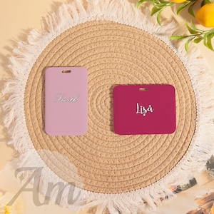 May include: Two rectangular luggage tags, one light pink and one magenta, each with a hole at the top. The light pink tag has the name "Smith" in cursive, and the magenta tag has the name "Lisa" in cursive. Both tags are on a woven placemat.