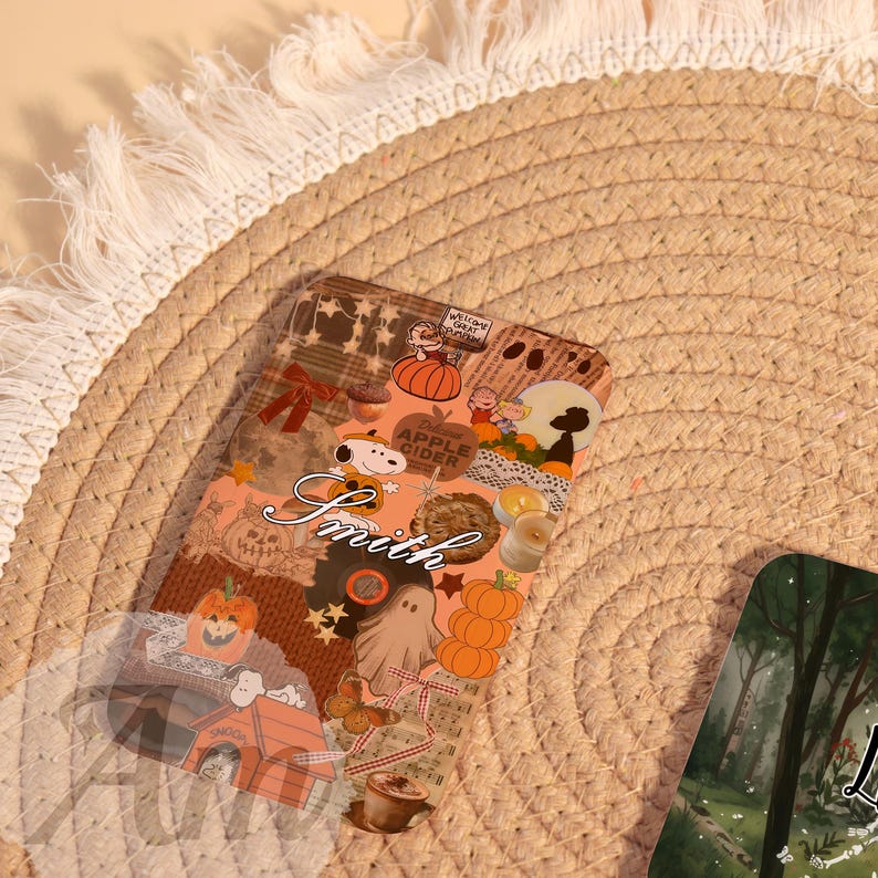 May include: A clear phone case with a collage of fall-themed images, including pumpkins, Snoopy, and the words "Apple Cider." The name "Smith" is written in cursive. The case is on a woven, beige surface.