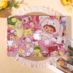 Op de afbeelding: Een roze laptophoes met een kleurrijke collage van cartoonfiguren. De hoes heeft een uitsparing voor het Apple-logo en is versierd met verschillende figuren in roze, groen, geel en wit. De hoes ligt op een ronde, geweven mat.