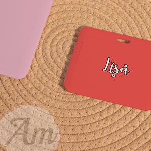 May include: A red rectangular card with the name "Lisa" in a cursive font. The card is on a woven surface, with a pink rectangular object in the upper left corner. The name "Am" is also visible.