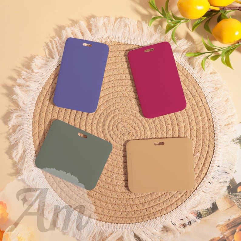 May include: Four rectangular ID badge holders in blue, magenta, olive green, and tan are arranged on a woven placemat. Each holder has a small hole at the top. The placemat is fringed with white. Lemons and greenery are in the background.