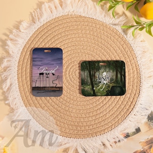 May include: Two rectangular luggage tags. One features a purple and gray night scene with the name "Smith" and a crow. The other tag displays a forest scene with the name "Lisa" and a skeleton. Both tags have a hole for attachment.