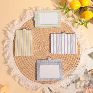 May include: Four ID badge holders with colourful striped patterns. The holders are rectangular with a clear window for displaying identification. They are arranged on a woven placemat with a fringed edge, with lemons and greenery in the background.