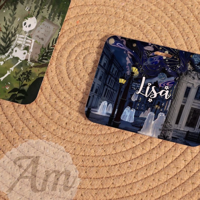 May include: Two decorative cards with illustrations. One features a skeleton in a graveyard setting. The other card has a night scene with ghosts, streetlights, and the name "Lisa" in script. The cards are on a woven surface.