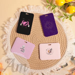 May include: Four rectangular cardholders with unique designs. One black cardholder features a pink crystal ball graphic. A purple cardholder displays a crescent moon and stars. Two pink cardholders have a skeleton reading and a witch graphic.