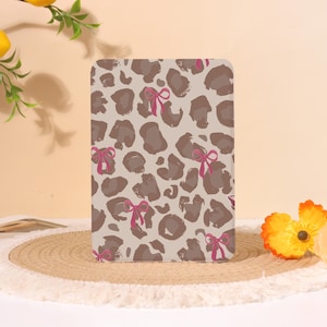 Op de afbeelding: Een rechthoekige tablethoes met een beige achtergrond, een bruin luipaardpatroon en kleine, felroze strikjes verspreid over het oppervlak. De hoes heeft afgeronde hoeken en een gladde, platte afwerking aan de voorzijde.