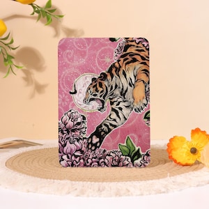 Peut inclure: Un objet décoratif rectangulaire avec une illustration détaillée d'un tigre bondissant en avant. Le tigre est orange et noir sur un fond rose avec des éléments floraux et lunaires. L'objet est entouré de fleurs roses et de feuilles vertes.