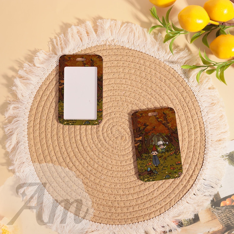May include: Two rectangular ID badge holders with a forest scene design. One holder has a white insert, the other features the full illustration. The holders are brown with a small hole at the top for a lanyard.