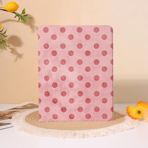 Pode incluir: Uma capa retangular para tablet com um fundo rosa claro e um padrão repetitivo de bolinhas rosa escuro. O estojo possui cantos arredondados e uma superfície plana e lisa, posicionada na vertical com um design consistente.