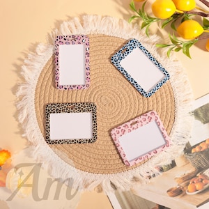 Può includere: Quattro porta badge rettangolari con motivi leopardati rosa, marroni e blu. Ogni supporto ha uno spazio rettangolare bianco per una carta d'identità. I supporti sono disposti su un sottobicchiere rotondo e tessuto.