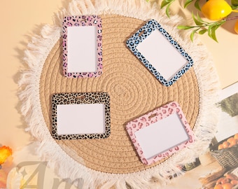 May include: Four rectangular ID badge holders with leopard print designs in pink, brown, and blue. Each holder has a white rectangular space for an ID card. The holders are arranged on a round, woven placemat.
