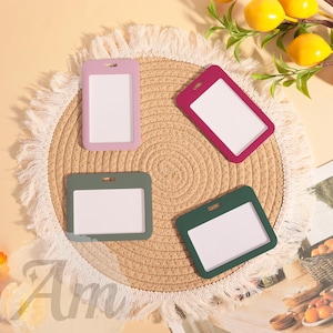 May include: Four colorful ID badge holders in pink, burgundy, and two shades of green, each with a white insert. The holders are arranged on a round, woven placemat with a fringed edge. Lemons and greenery are in the background.