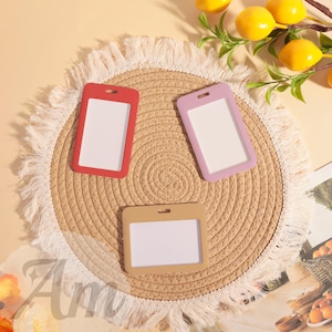 May include: Three ID badge holders in red, lavender, and tan, each with a white insert, are arranged on a round, woven placemat with a fringed edge. The background includes a sprig of yellow lemons.