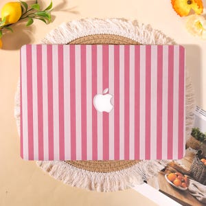 May include: A pink and white striped laptop cover with the Apple logo cutout. The cover is on a round, woven placemat with a fringed edge. The background includes lemons, flowers, and a basket of fruit.