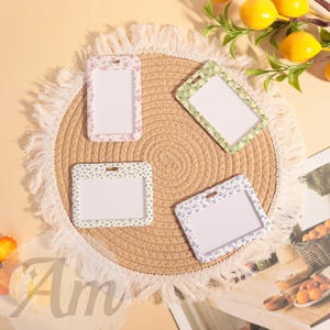May include: Four floral-patterned ID badge holders in pink, green, blue, and white. The holders are rectangular with a clear window and a small hole at the top. They are arranged on a round, woven mat.