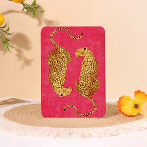 Pode incluir: Um item decorativo retangular rosa choque com dois leopardos estilizados. Os leopardos são retratados em amarelo dourado com manchas pretas. O fundo tem uma aparência texturizada. O item é colocado em uma superfície bege.