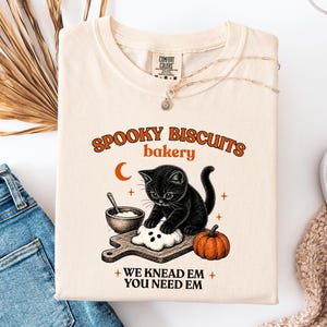 Pode incluir: Uma t-shirt de cor creme com um gráfico de um gatinho preto a amassar massa numa tábua de madeira. O texto diz SPOOKY BISCUITS bakery e WE KNEAD EM YOU NEED EM. O design inclui uma pequena taça, uma lua crescente e uma abóbora laranja.