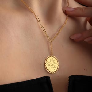 May include: A gold necklace with a circular pendant featuring the letters "S K B T E" arranged in a circle.