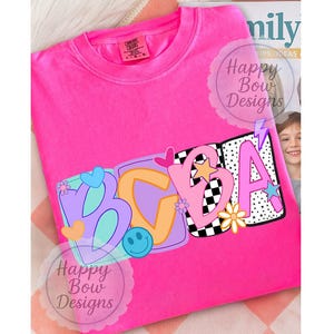 Op de afbeelding: Een felroze t-shirt met de letters "BCGA" in een kleurrijk, speels ontwerp. De letters zijn versierd met verschillende patronen, waaronder een dambord, sterren en bloemen. Het shirt heeft een ronde hals en korte mouwen.