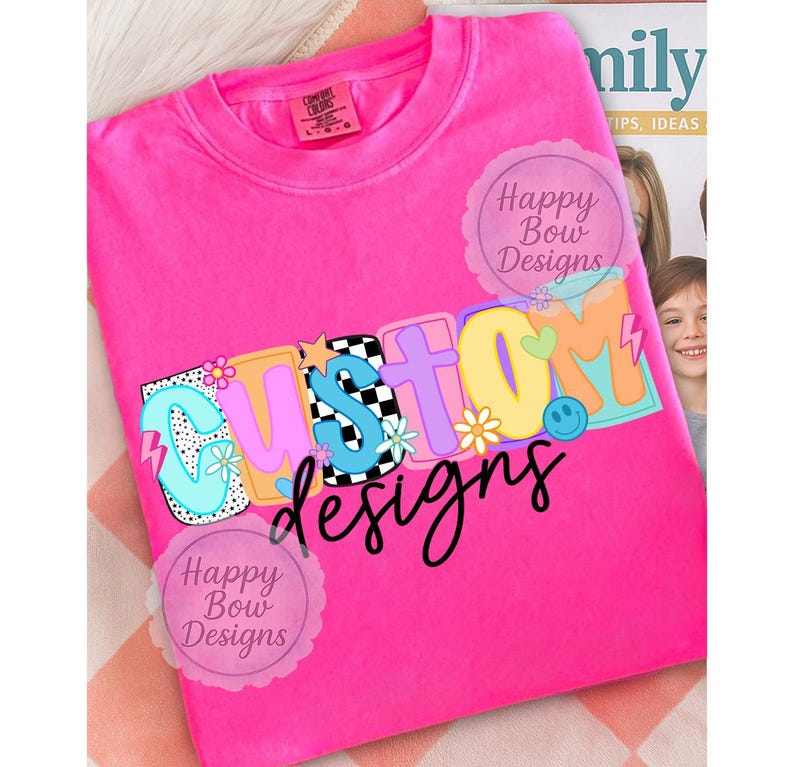 Pu&ograve; includere: Una maglietta rosa acceso con la scritta "CUSTOM" in lettere colorate e giocose. La parola "designs" &egrave; scritta in corsivo nero sotto. Il design include fiori, una faccina sorridente e un motivo a scacchi.