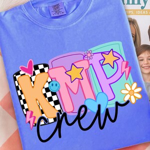 Pu&ograve; includere: Una maglietta blu pervinca con una grafica colorata che dice "KMP crew". La grafica include una "K" a scacchi bianchi e neri, una "M" rosa e una "P" viola, insieme a stelle, cuori e una faccina sorridente.