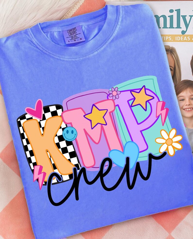 Pu&ograve; includere: Una maglietta blu pervinca con una grafica colorata che dice "KMP crew". La grafica include una "K" a scacchi bianchi e neri, una "M" rosa e una "P" viola, insieme a stelle, cuori e una faccina sorridente.