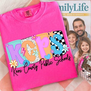 Pu&ograve; includere: Una maglietta rosa acceso con la scritta "KCPS Knox County Public Schools" in un design retr&ograve; colorato. Il design include fiori, stelle e un motivo a scacchiera. La maglietta &egrave; piegata su una superficie a fantasia.