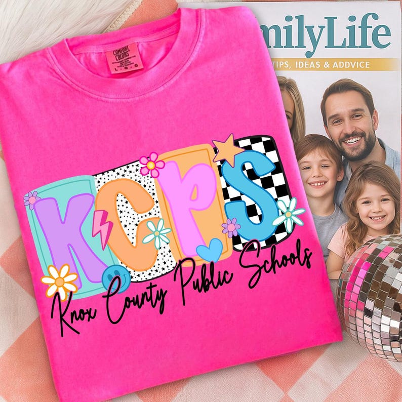 Pu&ograve; includere: Una maglietta rosa acceso con la scritta "KCPS Knox County Public Schools" in un design retr&ograve; colorato. Il design include fiori, stelle e un motivo a scacchiera. La maglietta &egrave; piegata su una superficie a fantasia.