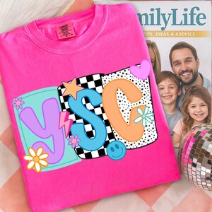 Pu&ograve; includere: Una felpa rosa acceso girocollo con un design grafico colorato. Il design include le lettere "YSC" in vari colori, insieme a stelle, fiori, un fulmine e una faccina sorridente. La felpa &egrave; esposta su una superficie a fantasia.
