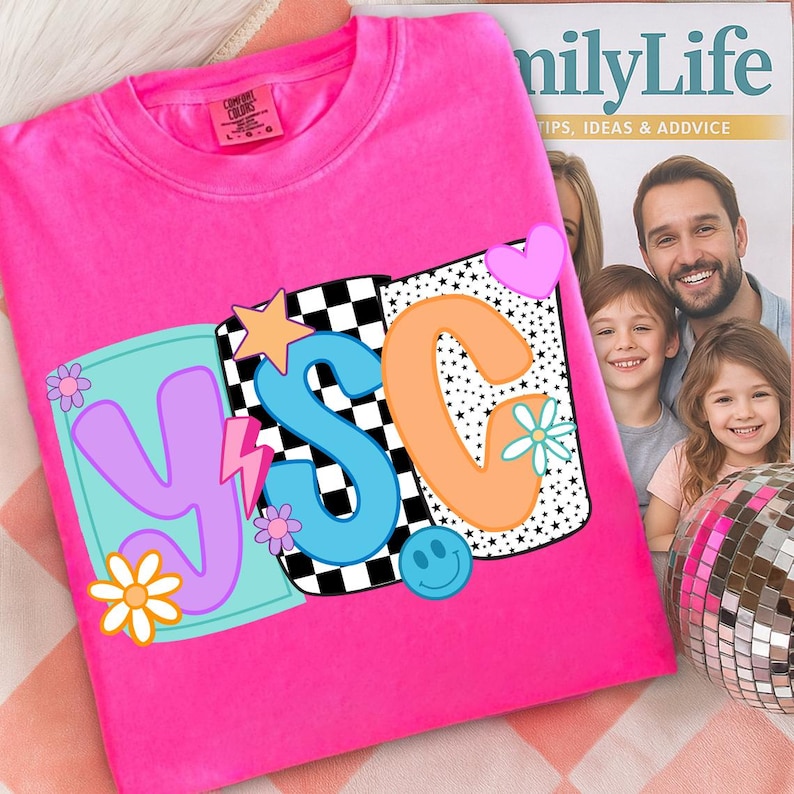 Pu&ograve; includere: Una felpa rosa acceso girocollo con un design grafico colorato. Il design include le lettere "YSC" in vari colori, insieme a stelle, fiori, un fulmine e una faccina sorridente. La felpa &egrave; esposta su una superficie a fantasia.