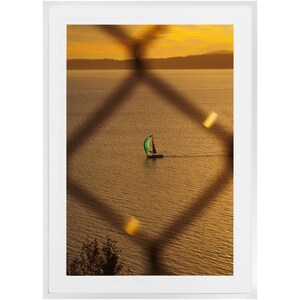 May include: A sailboat with a green sail is seen in the distance on a body of water. The water is a golden colour and the sky is a light orange. The scene is framed by a chain link fence.