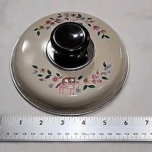 May include: Cream-colored pot lid with a black knob and a decorative floral and animal design. The design includes a swan, flowers, and a cat riding a donkey. The lid has a silver rim.