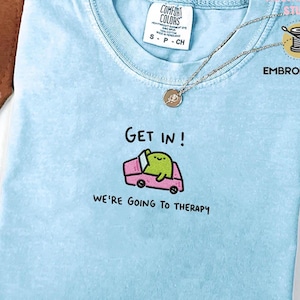 May include: Light blue t-shirt with the word "CHAMBRAY" in the upper left corner. The shirt features a graphic of a green creature in a pink car with the text "GET IN! WE'RE GOING TO THERAPY". A silver necklace is draped on the shirt.