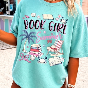 May include: A light turquoise t-shirt featuring the text "BOOK GIRL Summer" in black and pink. The graphic includes illustrations of a stack of books, a beach chair, a palm tree, crabs, seashells, starfish, and pink bows scattered across the front.