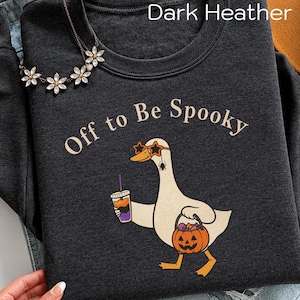 May include: A dark heather gray crewneck sweatshirt featuring a graphic of a white duck wearing star-shaped orange sunglasses, holding a drink and a jack-o'-lantern candy bucket. The text reads Off to Be Spooky in a light beige font.