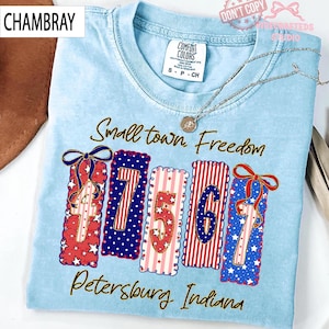 May include: Light blue t-shirt with the words "Small town Freedom" and "Petersburg Indiana" in script. The shirt features decorative rectangles with the numbers 4, 7, 5, 6, and 7 in red, white, and blue patterns. A silver necklace is draped across the shirt.
