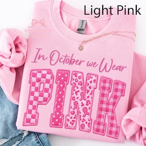 May include: A light pink crewneck sweatshirt featuring the text In October we Wear PINK in a bold font. The letters are filled with various patterns including checkers, polka dots, leopard print with breast cancer ribbons, and plaid.