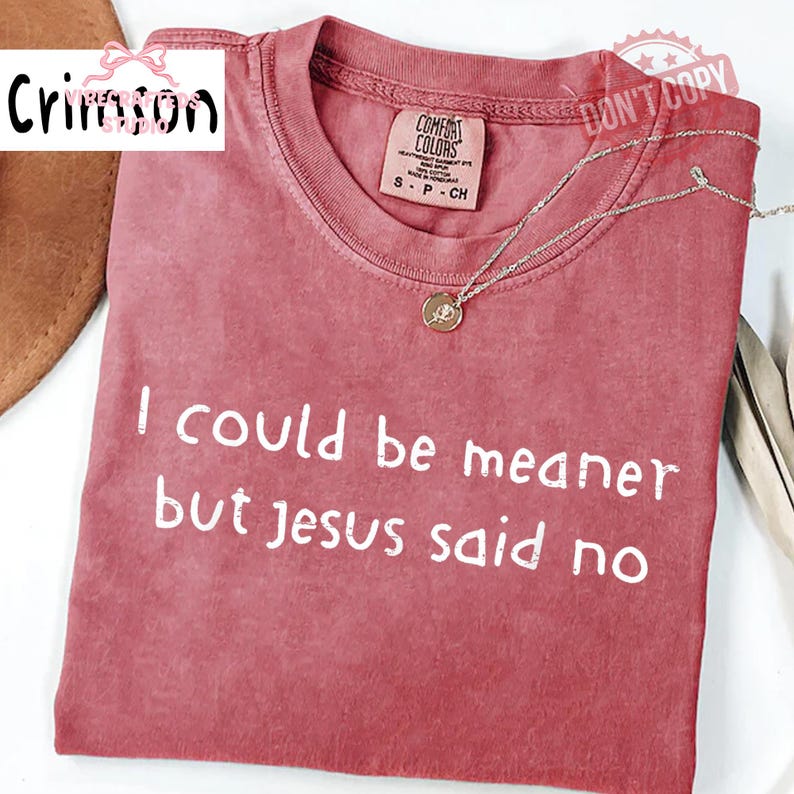 May include: A dusty rose-colored t-shirt with the text "I could be meaner but Jesus said no" in white. The shirt has a crew neck and short sleeves. A silver necklace with a small pendant rests on the shirt.