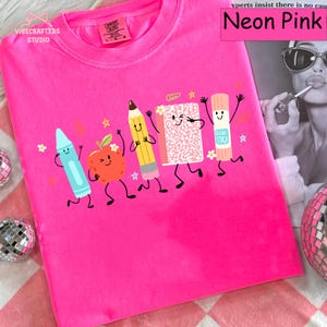 Puede incluir: Una camiseta de manga corta color rosa ne&oacute;n con un estampado gr&aacute;fico de cinco &uacute;tiles escolares antropom&oacute;rficos: un cray&oacute;n azul, una manzana roja, un l&aacute;piz amarillo, un cuaderno rosa y un pegamento en barra, todos con caras sonrientes.