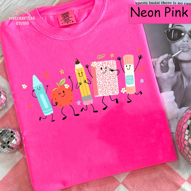 Puede incluir: Una camiseta de manga corta color rosa ne&oacute;n con un estampado gr&aacute;fico de cinco &uacute;tiles escolares antropom&oacute;rficos: un cray&oacute;n azul, una manzana roja, un l&aacute;piz amarillo, un cuaderno rosa y un pegamento en barra, todos con caras sonrientes.