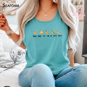 May include: A teal sleeveless tank top featuring a graphic of five small hot dogs in a row above the black text "bad day to be a hot dog". The shirt has a round neckline and is made of a solid-colored fabric, showcasing a casual design.