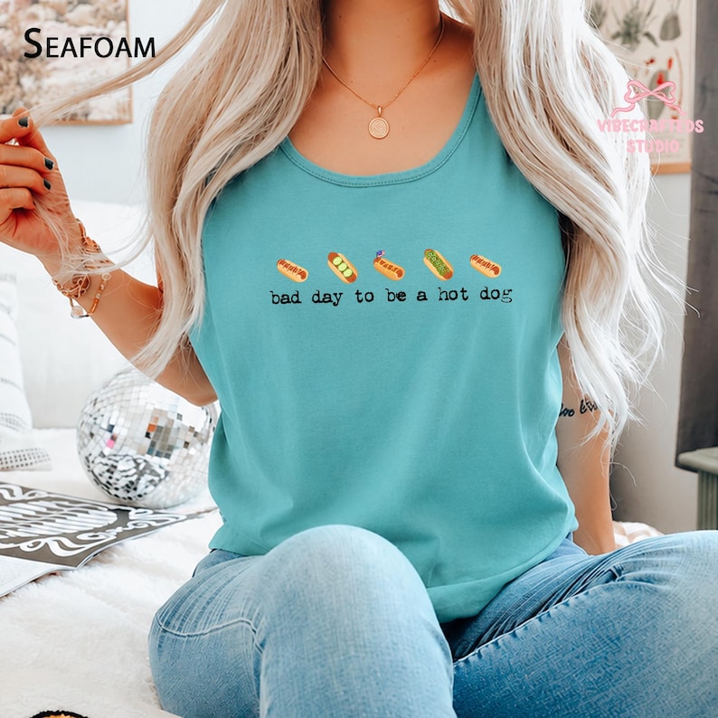 May include: A teal sleeveless tank top featuring a graphic of five small hot dogs in a row above the black text "bad day to be a hot dog". The shirt has a round neckline and is made of a solid-colored fabric, showcasing a casual design.