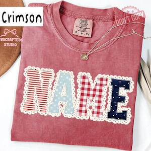 May include: Crimson-colored t-shirt with the word "NAME" appliqued across the front. The letters are in various patterns: red stripes, light blue polka dots, red gingham, and navy blue polka dots. A silver necklace with a pendant is draped on the shirt.