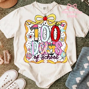 Puede incluir: Camiseta color crema con un gr&aacute;fico colorido que celebra 100 d&iacute;as de escuela. El dise&ntilde;o incluye un gran "100" con un l&aacute;piz, una manzana y las palabras "Days of School". La camiseta tiene cuello redondo y mangas cortas.