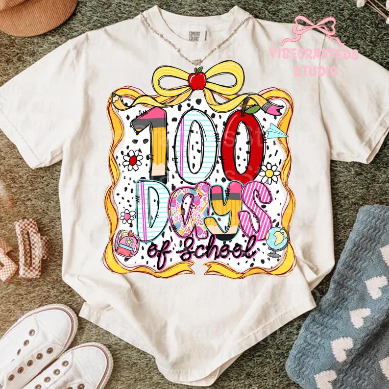 Puede incluir: Camiseta color crema con un gr&aacute;fico colorido que celebra 100 d&iacute;as de escuela. El dise&ntilde;o incluye un gran "100" con un l&aacute;piz, una manzana y las palabras "Days of School". La camiseta tiene cuello redondo y mangas cortas.
