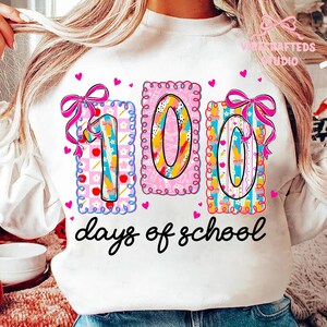 May include: White sweatshirt with a colorful "100 days of school" graphic. The design features the numbers "100" in decorative frames with bows and hearts. The text "days of school" is written in a cursive font.
