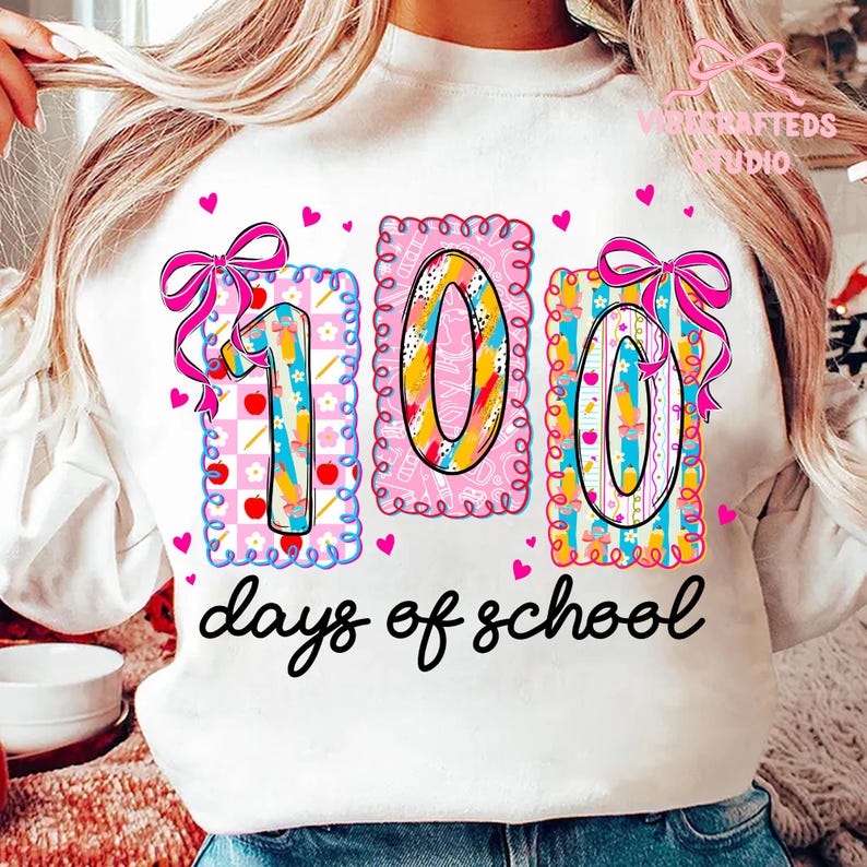 May include: White sweatshirt with a colorful "100 days of school" graphic. The design features the numbers "100" in decorative frames with bows and hearts. The text "days of school" is written in a cursive font.