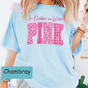 May include: A light blue short-sleeve t-shirt featuring the text In October we Wear PINK in pink lettering. The word PINK is displayed in large block letters with varied patterns including checkers, polka dots, leopard print, and plaid.